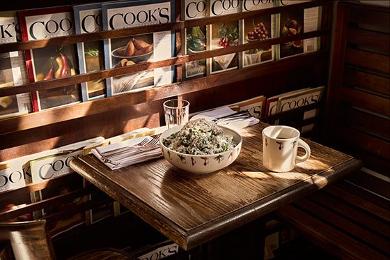 Dining table with a bowl of salad and a mug, surrounded by cookbooks on a wooden shelf.