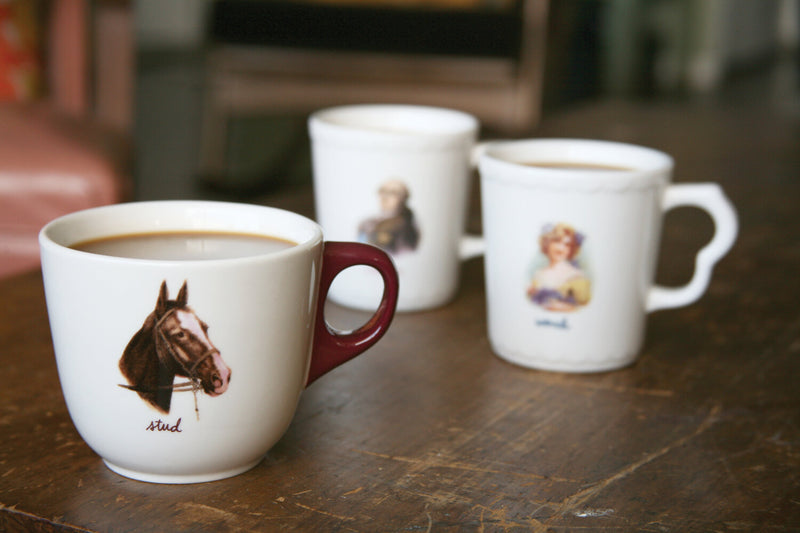 Three mugs on a wooden table featuring All In The Family designs, such as the Stud Mug with a horse design. 